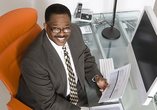 A mature businessman sitting at his desk holding papers and smiling up at the camera A mature businessman holding papers in his hand and sitting at his desk